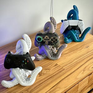 May include: Three octopus-shaped controller holders in white, gray, and blue. Each holder cradles a different game controller: a black Xbox controller, a gray controller, and a white and black PlayStation controller. The holders are on a wooden surface.