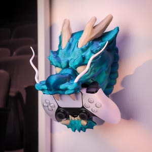 May include: A wall-mounted dragon head sculpture in a vibrant teal and blue gradient finish, featuring long white whiskers and beige horns. The dragon's open mouth is designed to hold a white and black wireless video game controller securely.