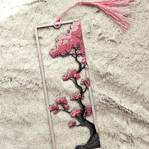May include: A pink rectangular bookmark featuring a cherry blossom tree design. The tree has a black trunk and branches with pink blossoms. A pink tassel hangs from the top. The bookmark is set against a soft, textured background.