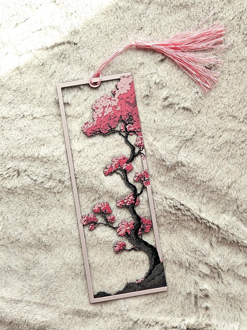 May include: A pink rectangular bookmark featuring a cherry blossom tree design. The tree has a black trunk and branches with pink blossoms. A pink tassel hangs from the top. The bookmark is set against a soft, textured background.