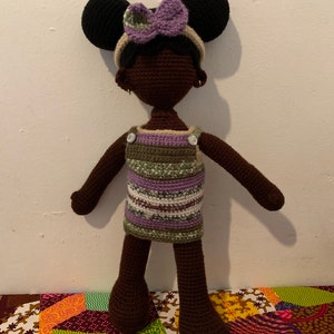 May include: A handmade crochet doll with dark brown skin, wearing a striped dress in shades of green, purple, and white. The doll has black hair styled with two large round buns and a purple bow. The doll is standing on a colorful patterned fabric.