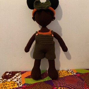 May include: A crocheted doll with brown skin, black hair, and green overalls. The doll is wearing a headband with a green bow. The doll is standing on a colorful patterned fabric.