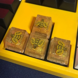 Puede incluir: Cuatro teclas amarillas con un diseño de abeja y purpurina. Las teclas están en un teclado amarillo.