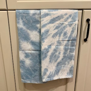 以下が含まれることがあります： 白のキャビネットドアに掛けられた、水色と白のタイダイ柄のキッチンタオル2枚。タオルは柔らかく、質感のある外観で、半分に折りたたまれています。キャビネットには黒いハンドルが付いています。