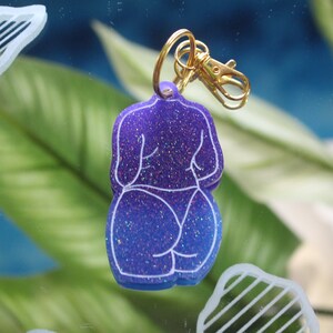 May include: A glittery purple and blue keychain in the shape of a figure's backside. The keychain has a gold-colored clasp and ring. The figure's outline is white.