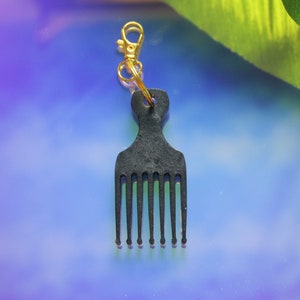 Puede incluir: Un llavero de peine afro negro con un mosquetón y un anillo dorados. El peine tiene siete púas y un mango con un agujero para el anillo. El fondo es un degradado de azul y morado.