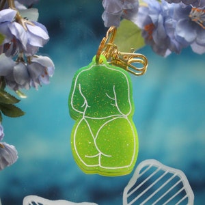 May include: A green glittery keychain in the shape of a person's silhouette, viewed from the back. The keychain has a gold-colored clasp and a white outline. The background is a blurred blue with purple flowers.