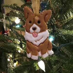 May include: A brown and white corgi dog ornament with a pink tongue, hanging from a gold ribbon. The ornament is on a Christmas tree with lights.