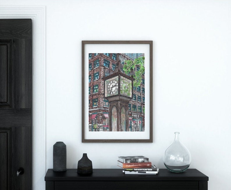 May include: Framed art print featuring a detailed watercolor illustration of a vintage street clock. The clock is brown with a glass face, set against a backdrop of brick buildings and greenery. The artwork is displayed on a white wall.
