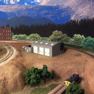 May include: A model train scene featuring a gray building with three garage doors, set against a backdrop of mountains and trees. A train with brown and yellow cars is on the tracks, with green trees and brown terrain.