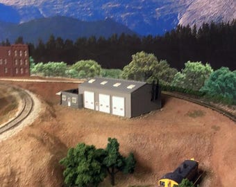 May include: A model train scene featuring a gray building with three garage doors, set against a backdrop of mountains and trees. A train with brown and yellow cars is on the tracks, with green trees and brown terrain.