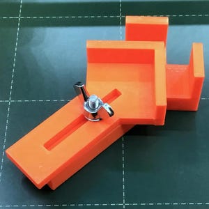 May include: Orange plastic corner clamp with a silver adjustable knob. The clamp is designed to hold two pieces of wood or other materials at a 90-degree angle.