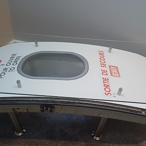 May include: A distinctive coffee table crafted from a recycled aircraft door. It has a clear glass top, a white door panel with red text, and metal legs. The door panel includes an oval window and is about 152 cm long.