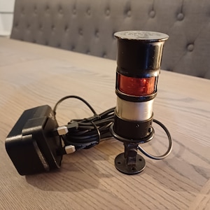 Puede incluir: Una lámpara náutica vintage negra y plateada con una lente roja, montada sobre una base circular. La lámpara está conectada a un adaptador de corriente negro a través de un cable negro. La lámpara está sobre una superficie de madera clara.