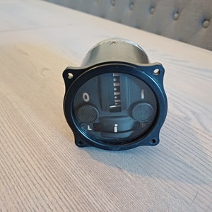 Puede incluir: Un instrumento circular de avión negro con un dial blanco y una aguja. El dial tiene marcas de 0 a 1 y las palabras "L" y "R" a cada lado de la aguja. El instrumento tiene dos botones negros a cada lado del dial.