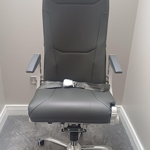 Peut inclure: Une chaise de bureau à dossier haut recouverte de similicuir gris foncé avec un coussin d'assise perforé. La chaise présente des accoudoirs argentés, une base chromée à cinq branches avec roulettes et une ceinture de sécurité grise.