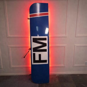 Op de afbeelding: Een hoog, blauw bord met de letters "FM" in zwart en wit. Het bord heeft een rood-witte streep aan de bovenkant en wordt verlicht met rode lichten. Het bord is aan een muur bevestigd.