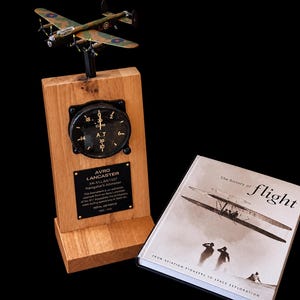 Op de afbeelding: Een houten displaystandaard met een vintage zwarte ronde hoogtemeter en een klein Avro Lancaster modelvliegtuig bovenop. Een plaquette beschrijft de geschiedenis van het instrument. Daarnaast ligt een boek getiteld The History of Flight.