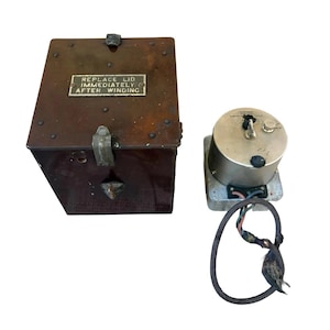 Könnte beinhalten: Eine Vintage-Holzkiste in Braun mit Metallbeschlägen und einem kleinen Metallschild mit der Aufschrift "REPLACE LID IMMEDIATELY AFTER WINDING". Daneben ein silberfarbenes mechanisches Gerät mit Zifferblatt und Drähten.