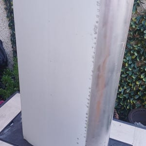 Pode incluir: Uma seção retangular de asa de avião feita de alumínio escovado com uma superfície lisa pintada em cinza claro em um lado. O componente apresenta uma borda de ataque curva fixada com uma fileira vertical de rebites metálicos visíveis.