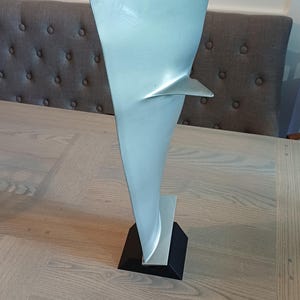 May include: A silver-colored metal turbine blade sculpture mounted on a solid black square base. The blade features a smooth, tapered vertical design with a small, triangular protrusion extending from the upper side of the main body.