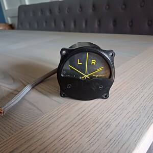 Op de afbeelding: Een zwarte ronde meter met een gele naald en markeringen. De meter heeft de letters "L" en "R" erop gedrukt. De meter is verbonden met een grijze draad.