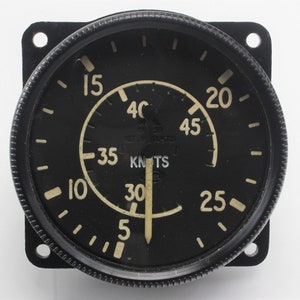 May include: A black and white aircraft instrument with a round face and a needle pointing to 30. The face has markings for knots and numbers from 5 to 45. The instrument has a black bezel and a silver ring around the face.