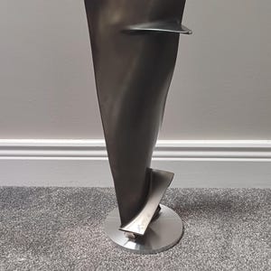 May include: A modern, abstract sculpture with a dark metallic finish. The piece features a curved, blade-like form rising from a circular base. A smaller wing-like element extends from the main body. The sculpture is displayed on a gray carpet.