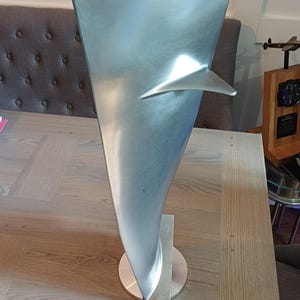 May include: A silver-colored metal turbine blade sculpture with a smooth, tapered, and curved design. The object features a small, triangular protrusion on one side and is mounted vertically on a circular base, showcasing a metallic finish.