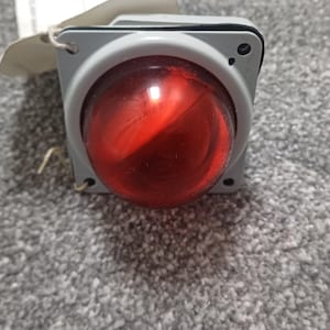 Puede incluir: Una luz de señalización industrial de forma cuadrada con una lente de cúpula roja translúcida prominente centrada en una carcasa gris claro. El dispositivo incluye cuatro orificios de montaje en las esquinas y una etiqueta de papel.