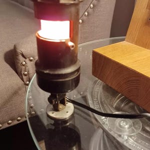 May include: A vintage-style lamp with a cylindrical, dark brown body and a bright, warm light emanating from within. The lamp sits on a clear glass table, with a metal base and a black power cord. The background includes a gray upholstered sofa.