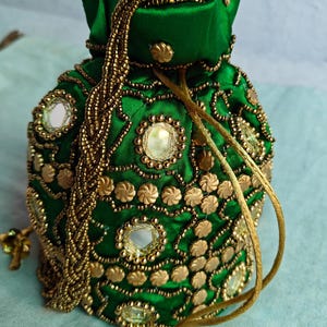 May include: A green satin drawstring bag, heavily embellished with gold-colored beads, sequins, and decorative accents. The bag features a drawstring closure and decorative tassels. The bag's design includes mirrored elements and floral-shaped embellishments.