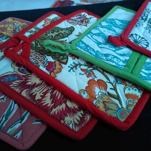 Könnte beinhalten: Vier farbenfrohe, gesteppte Topflappen mit floralen und geometrischen Mustern. Die Topflappen haben rote, grüne, blaue und weiße Akzente.