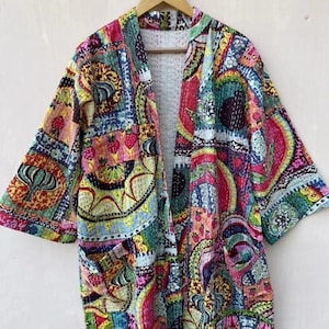 Könnte beinhalten: Ein farbenfroher Kimono-Stil-Bademantel mit offenem Schnitt und einem lebendigen Patchwork-Design. Der Bademantel weist eine Mischung aus Mustern und Farben auf, darunter Rosa, Gelb, Grün und Blau. Er hat lange Ärmel und zwei Vordertaschen und hängt an einem Holzbügel.