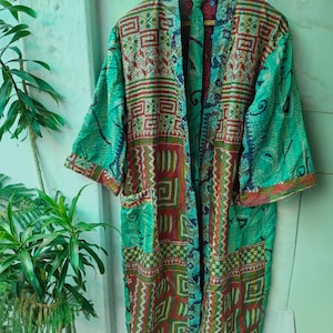 May include: A long, open-front kimono-style robe in shades of teal, red, and brown. The robe features a geometric pattern with a patchwork design. The sleeves are three-quarter length. The robe is hanging on a wooden hanger.