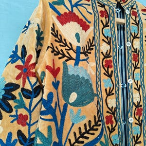 K&ouml;nnte beinhalten: Eine gelbe Samtjacke mit aufwendiger Blumenstickerei in Blau-, Rot- und Wei&szlig;t&ouml;nen. Die Jacke hat einen Stehkragen und einen Knopfverschluss.