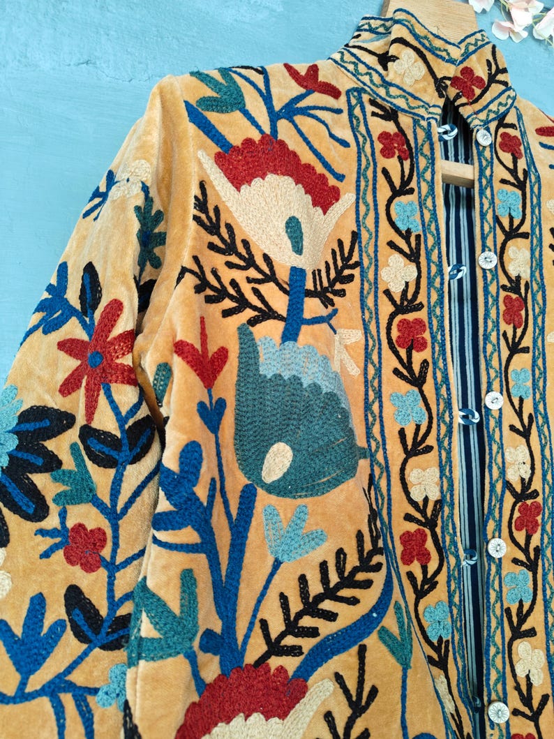 K&ouml;nnte beinhalten: Eine gelbe Samtjacke mit aufwendiger Blumenstickerei in Blau-, Rot- und Wei&szlig;t&ouml;nen. Die Jacke hat einen Stehkragen und einen Knopfverschluss.