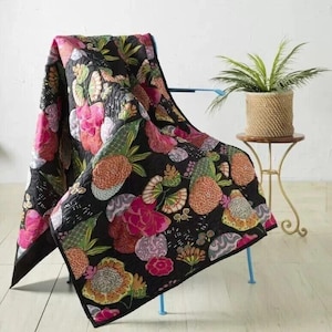Peut inclure: Une couverture noire ornée d'un motif floral aux couleurs vives, rose, orange et vert. La couverture est drapée sur une chaise bleue, mettant en valeur son design complexe et sa texture matelassée. Un pot de plante est également visible.