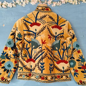 K&ouml;nnte beinhalten: Eine senfgelbe Wildlederjacke mit aufwendiger Blumenstickerei in Blau-, Rot- und Cremet&ouml;nen. Die Stickerei zeigt stilisierte Blumen und Bl&auml;tter.