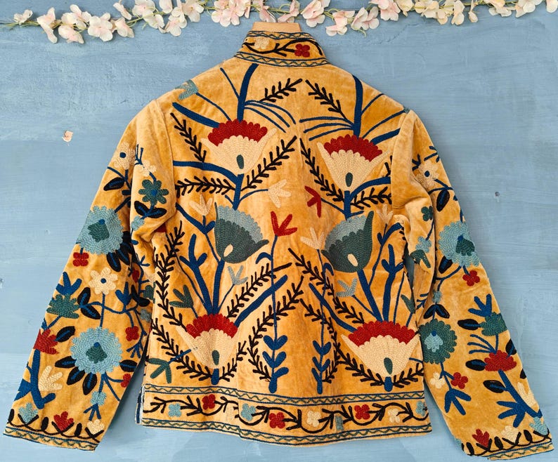 K&ouml;nnte beinhalten: Eine senfgelbe Wildlederjacke mit aufwendiger Blumenstickerei in Blau-, Rot- und Cremet&ouml;nen. Die Stickerei zeigt stilisierte Blumen und Bl&auml;tter.