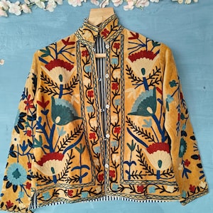 K&ouml;nnte beinhalten: Eine gelbe Samtjacke mit aufwendigen Blumenstickereien in Blau-, Rot- und Gr&uuml;nt&ouml;nen. Die Jacke hat einen hohen Kragen und einen Knopfverschluss.