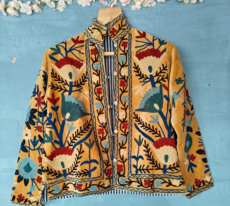 K&ouml;nnte beinhalten: Eine gelbe Samtjacke mit aufwendigen Blumenstickereien in Blau-, Rot- und Gr&uuml;nt&ouml;nen. Die Jacke hat einen hohen Kragen und einen Knopfverschluss.