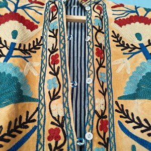 K&ouml;nnte beinhalten: Eine gelbe Wildlederjacke mit aufwendigen Blumenstickereien in Blau-, Rot- und Gr&uuml;nt&ouml;nen. Die Jacke hat einen Stehkragen und einen Knopfverschluss.