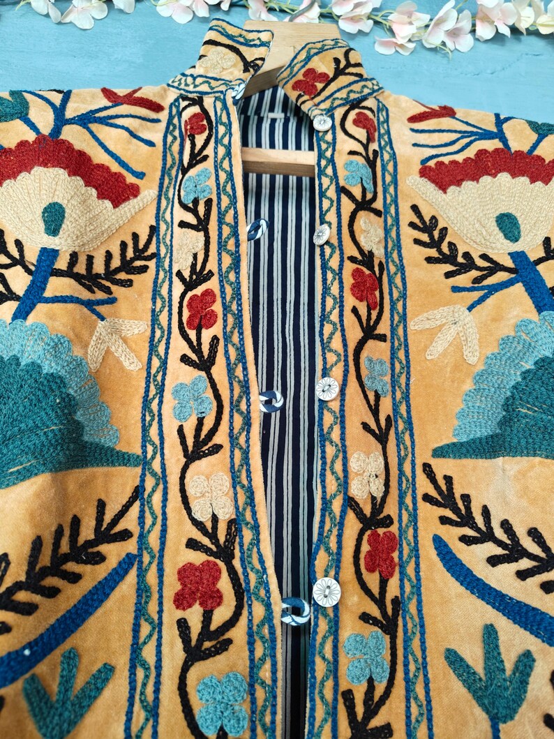 K&ouml;nnte beinhalten: Eine gelbe Wildlederjacke mit aufwendigen Blumenstickereien in Blau-, Rot- und Gr&uuml;nt&ouml;nen. Die Jacke hat einen Stehkragen und einen Knopfverschluss.