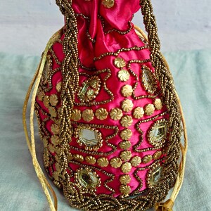 Op de afbeelding: Een roze trekkoordtas met gouden en spiegelende versieringen. De tas is versierd met een bloemenpatroon en heeft een kwast.