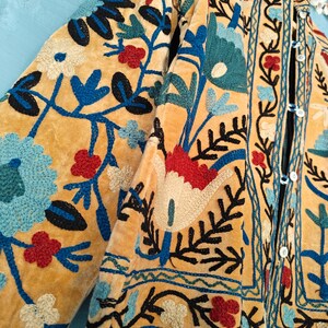 K&ouml;nnte beinhalten: Eine gelbe Samtjacke mit aufwendiger Blumenstickerei in Blau-, Rot- und Gr&uuml;nt&ouml;nen. Die Jacke hat einen Knopfverschluss und einen Stehkragen.