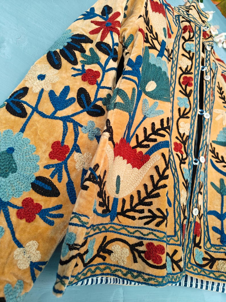 K&ouml;nnte beinhalten: Eine gelbe Samtjacke mit aufwendiger Blumenstickerei in Blau-, Rot- und Gr&uuml;nt&ouml;nen. Die Jacke hat einen Knopfverschluss und einen Stehkragen.