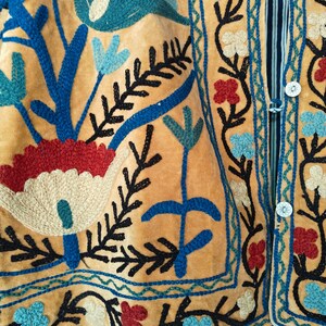 K&ouml;nnte beinhalten: Nahaufnahme eines gelben Stoffes mit aufwendigen Stickereien in blauer, roter und schwarzer Farbe. Die Stickerei zeigt florale Muster und geometrische Designs.