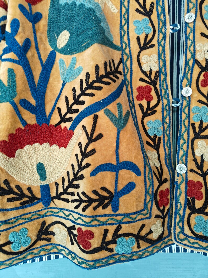 K&ouml;nnte beinhalten: Nahaufnahme eines gelben Stoffes mit aufwendigen Stickereien in blauer, roter und schwarzer Farbe. Die Stickerei zeigt florale Muster und geometrische Designs.