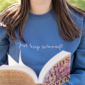 May include: A blue sweatshirt with the text "just keep swimming" embroidered in white. The sweatshirt is being worn by a person holding a book titled "Friends, Lovers, and the Big Terrible Thing" by  Jenny Lawson.